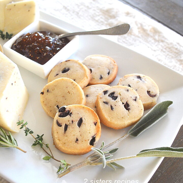 A white platter filled with shortbread biscuits studded with black olives, served with fig jam as an appetizer.