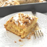 A slice of coffee cake with white icing drizzled on top, and served on a white plate with a fork.