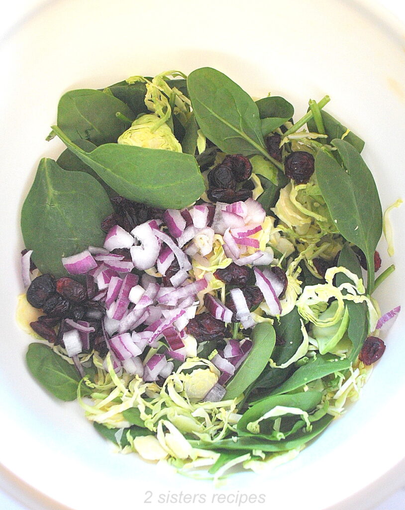 A white mixing bowl filled with fresh baby spinach, dried cranberries, shaved Brussels sprouts, chopped apple, and chopped red onion.