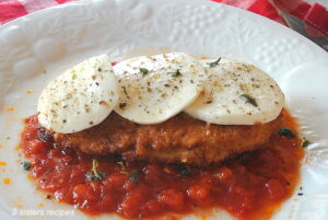 A white plate with tomato sauce, a fried chicken cutlet on top and sliced mozzarella over the chicken cutlet.