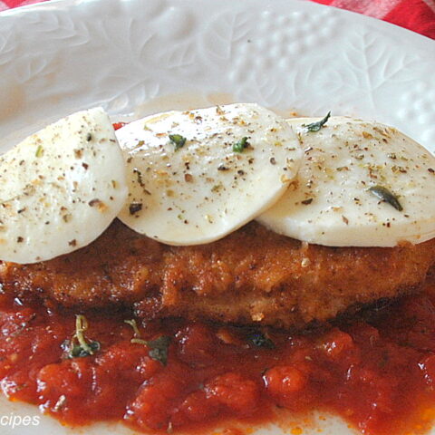 A white plate with tomato sauce, a fried chicken cutlet on top and sliced mozzarella over the chicken cutlet.