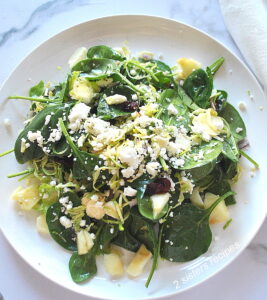A white dinner plate, served a salad, with spinach, chopped apple, shaved brussels sprouts, topped with crumbled cheese.
