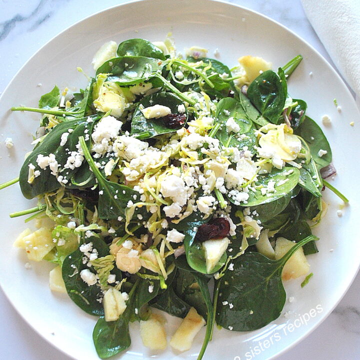 A white dinner plate, served a salad, with spinach, chopped apple, shaved brussels sprouts, topped with crumbled cheese.