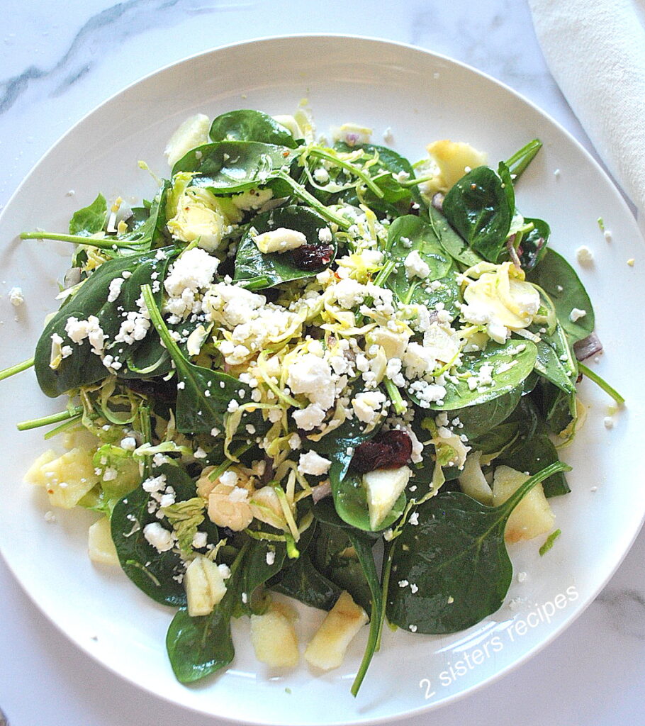 A white dinner plate, served a salad, with spinach, chopped apple, shaved brussels sprouts, topped with crumbled cheese.
