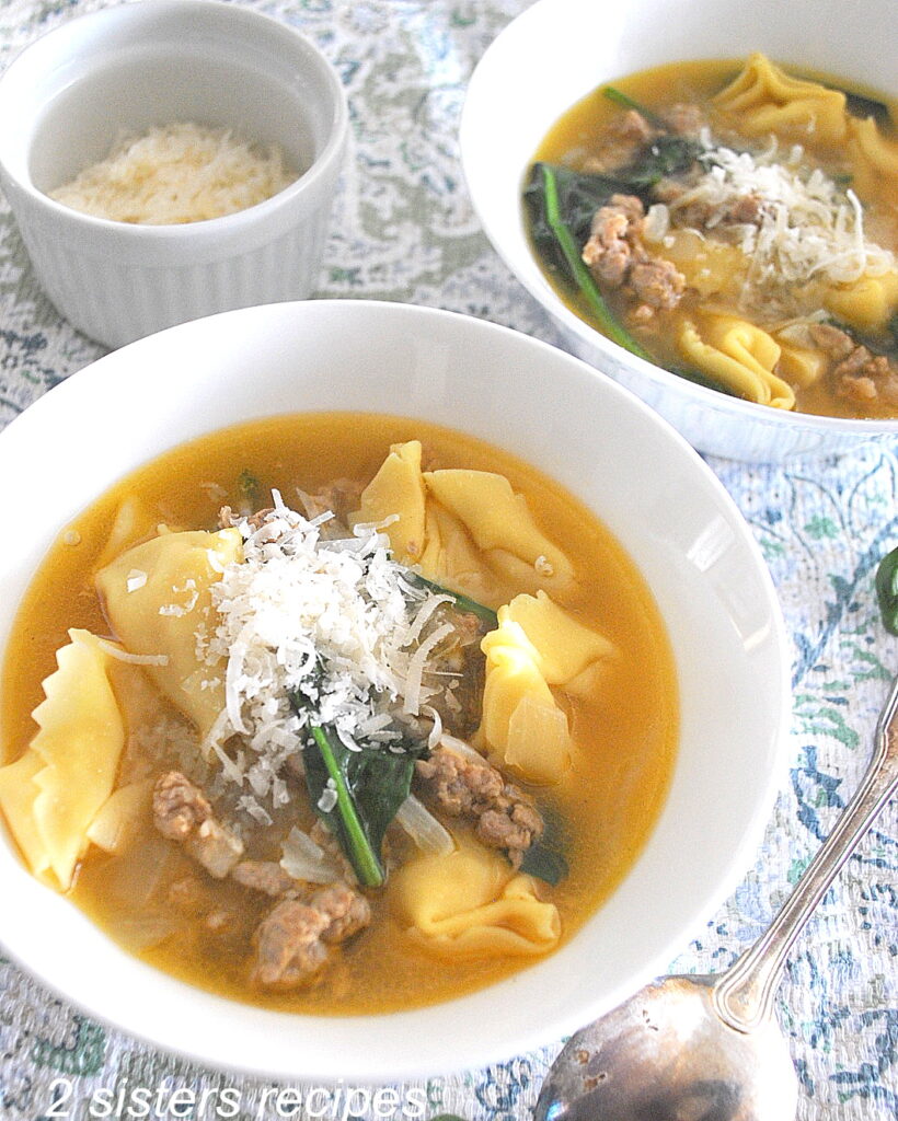 Two white bowls filled with Tortellini, spinach and crumbled sausage soup. Topped with grated cheese.