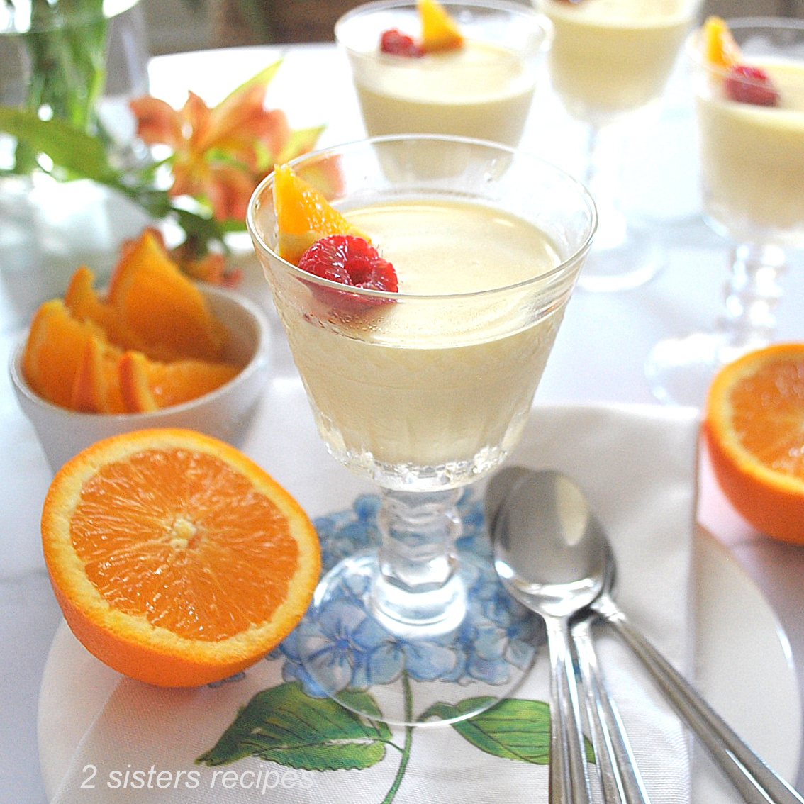 A stemware glass filled with an orange pudding, topped with a raspberry and orange wedge, plus sliced oranges on the side.