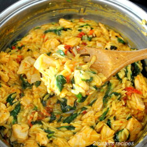 A skillet with cooked orzo pasta, chicken bites, wilted spinach and sun-dried tomatoes.