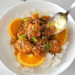 A white bowl filled with a bed of white rice, topped with orange pieces of cooked chicken, sliced oranges on the side. And topped with sesame seeds and green onions.