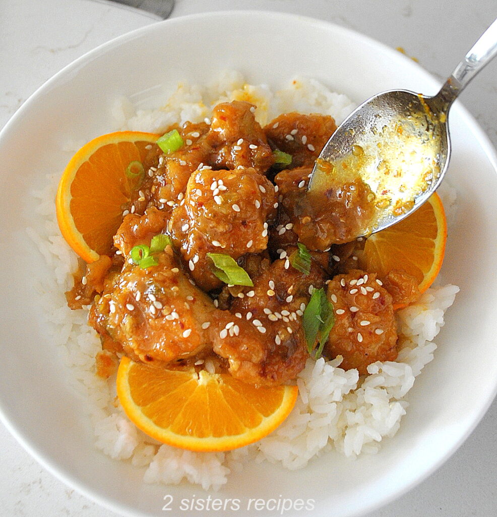 A white bowl filled with a bed of white rice, topped with orange pieces of cooked chicken, sliced oranges on the side. And topped with sesame seeds and green onions.