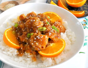 A white bowl filled with white rice, topped with orange pieces of chicken, sliced oranges, chopped scallions and sesame seeds