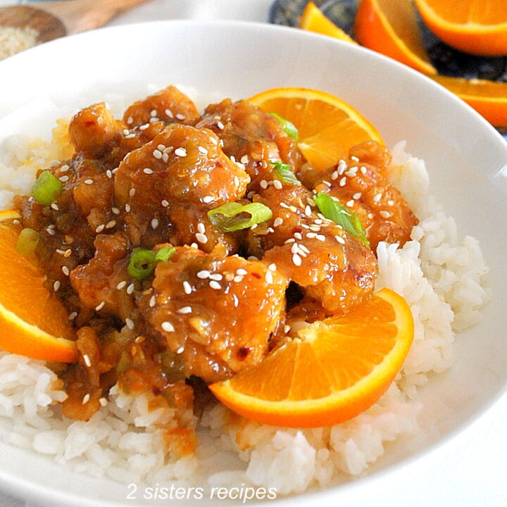 A white bowl filled with white rice, topped with orange pieces of chicken, sliced oranges, chopped scallions and sesame seeds