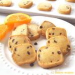 A white plate with small square orange chocolate chip shortbread cookies with slices of orange on the side..
