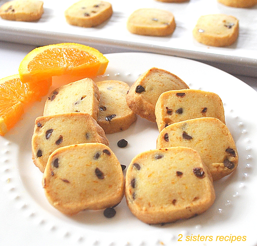 A white plate with small square orange chocolate chip shortbread cookies with slices of orange on the side..