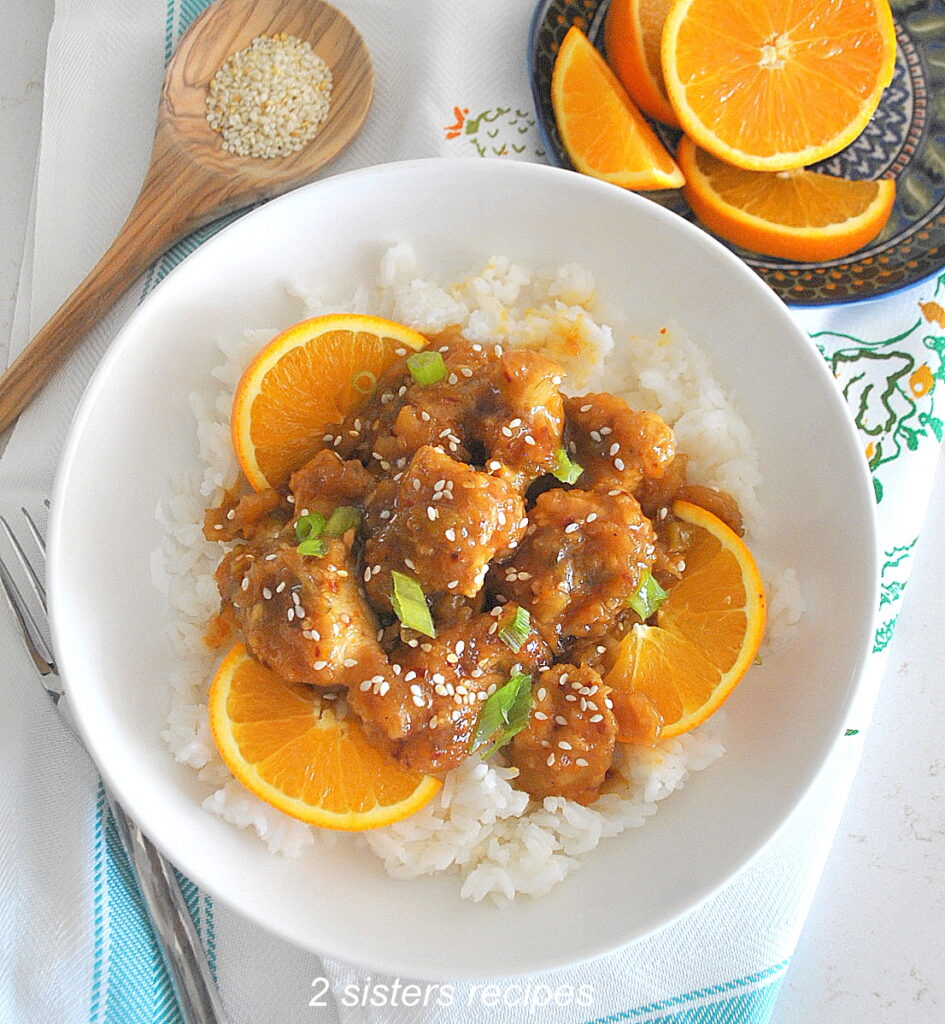 A white bowl filled witth Orange chicken over a bed of white rice. with sliced oranges as garnish.