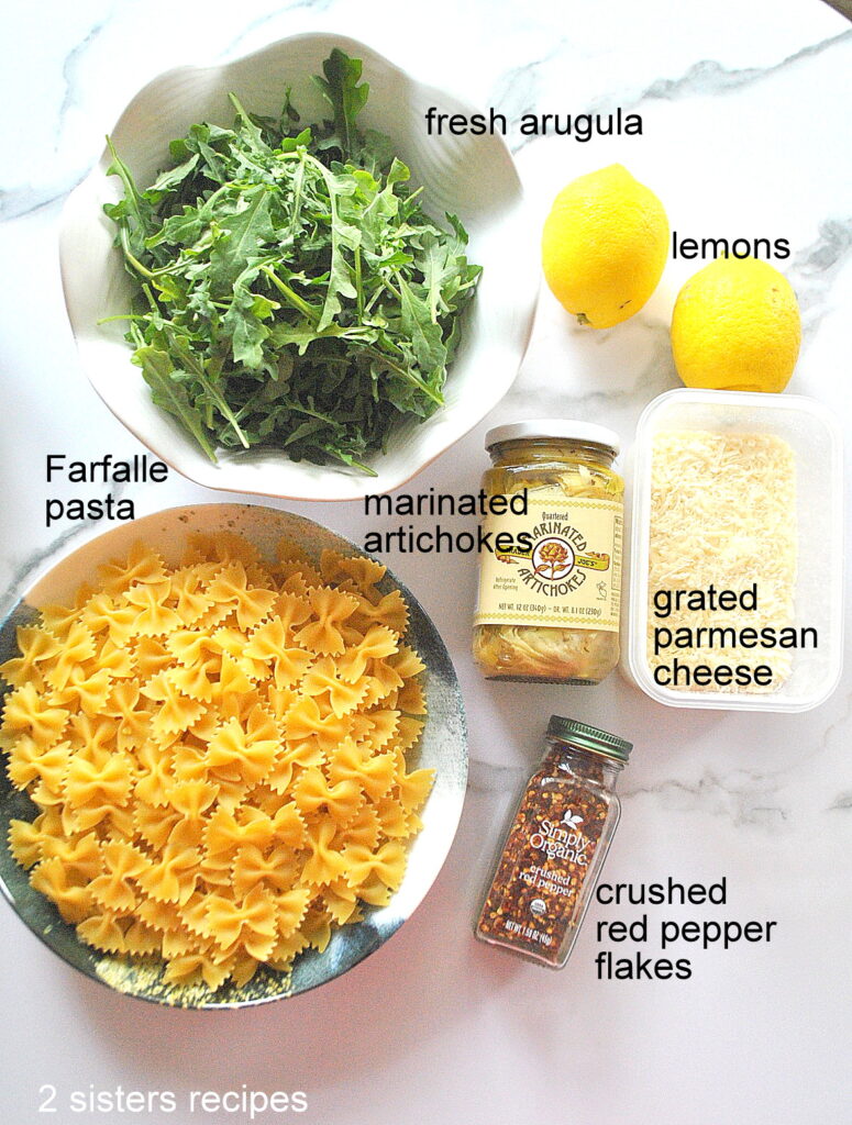 a bowl of fresh arugula, 2 lemons, a bowl of farfalle pasta, jar of artichokes, container of grated cheese and a bottle of red pepper flakes.