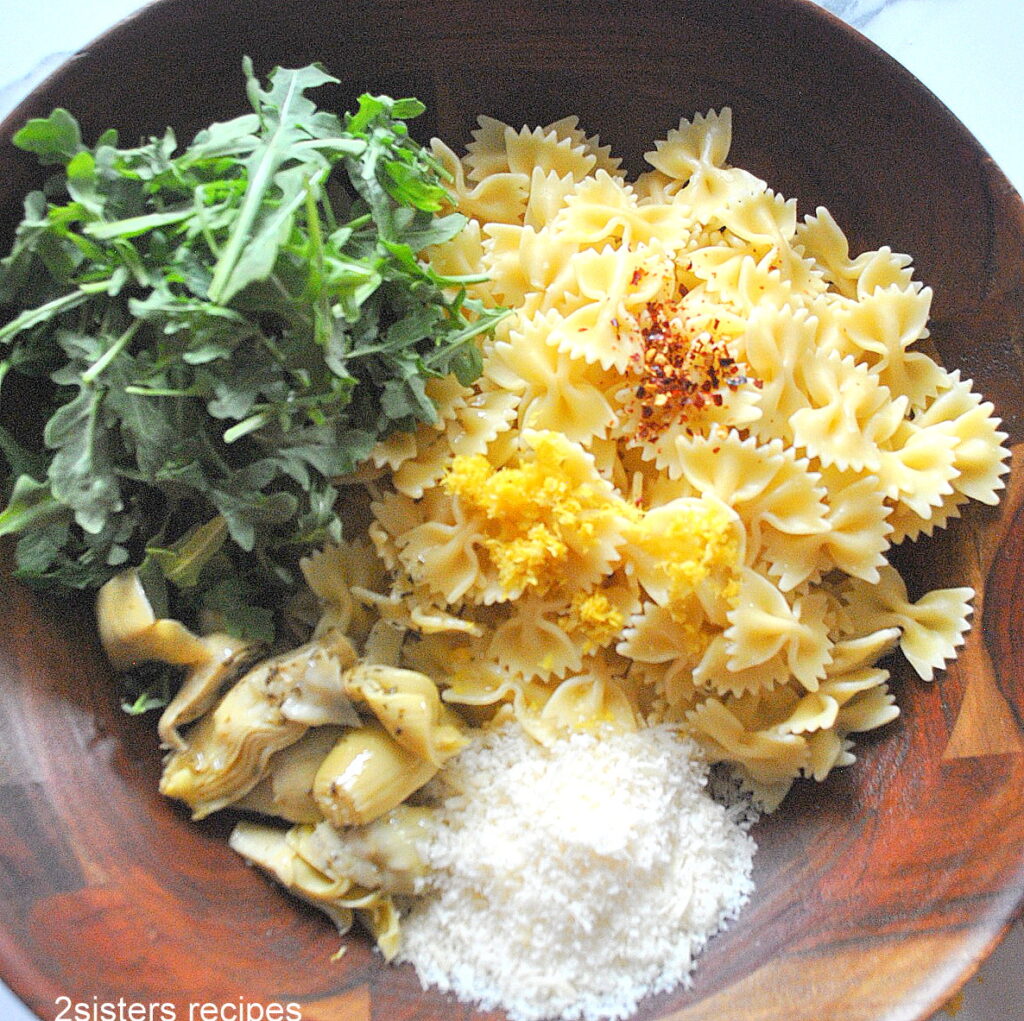 A wooden salad bowl with cooked farfalle pasta, fresh arugula, artichoke hearts, grated cheese and lemon zest.