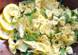 A wooden salad bowl filled with farfalle pasta, arugula and artichokes with slices of lemon on the side.