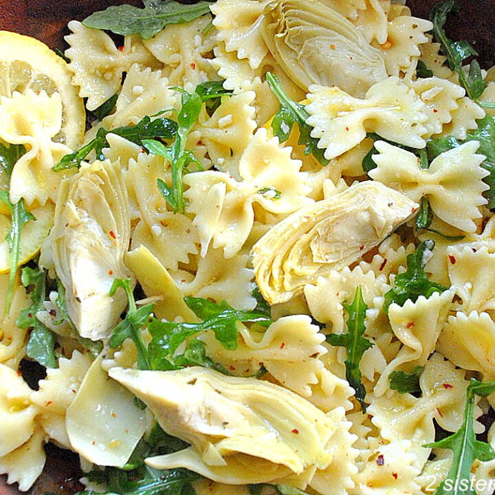 A wooden salad bowl filled with farfalle pasta, arugula and artichokes with slices of lemon on the side.