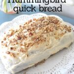 A hunningbird quick bread topped with white frosting and crushed pecans is served on a white doily.