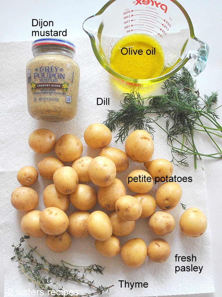the ingredients are laid on the counter. A jar of Dijon, glass cup or olive oil, fresh herbs, and small golden potatoes.
