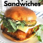 A fish sandwich topped with pickle, arugula, and cheese served on a white plate.