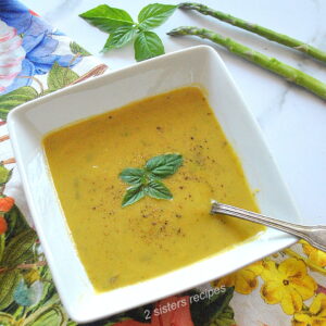 A square white bowl filled with a green colored pureed soup with a spoon in it.