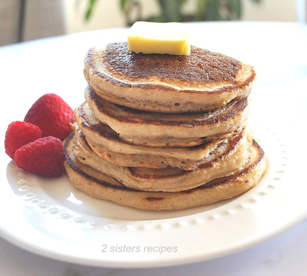 A white plate filled with a pile of eggless pancakes topped with a slice of butter and fresh strawberries on the side.