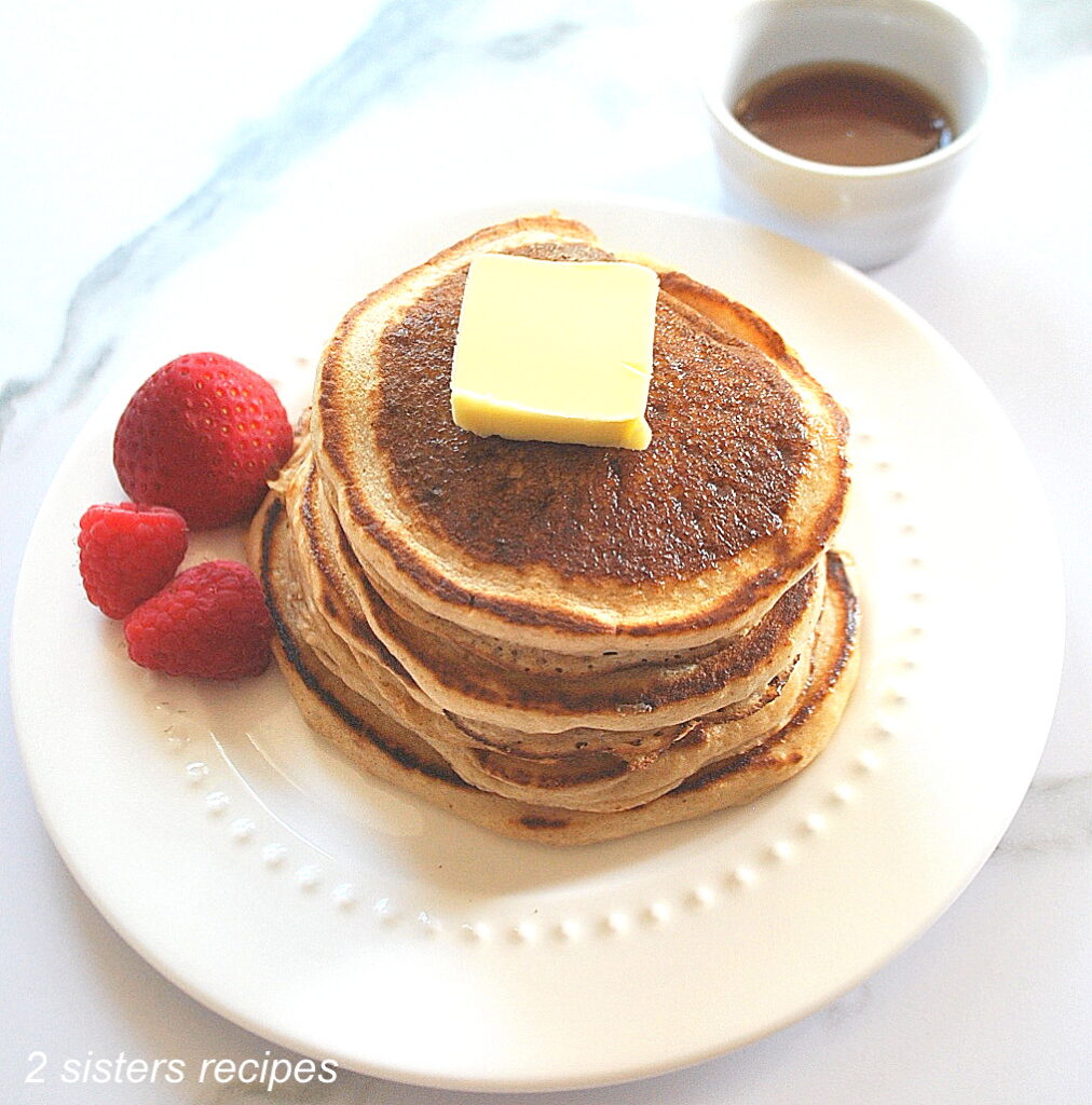 A white plate with a pile of pancakes, fresh fruit on the side. 