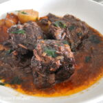 Three boneless short ribs with sauce served in a white plate with some veggies.