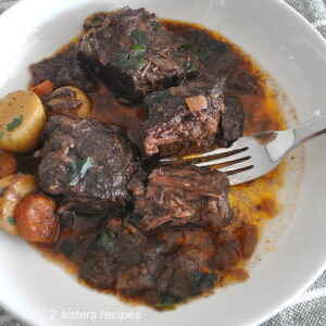 Three boneless short ribs in a white plate with some small potatoes, and carrots with a fork.