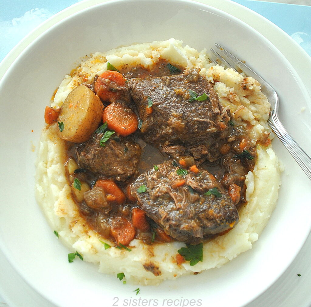 A white plate with a layer of mashed potatoes and boneless short ribs served on top.