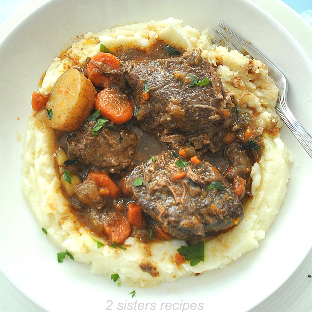A white plate filled with a layer of mashed potatoes topped with boneless short ribs and gravy.