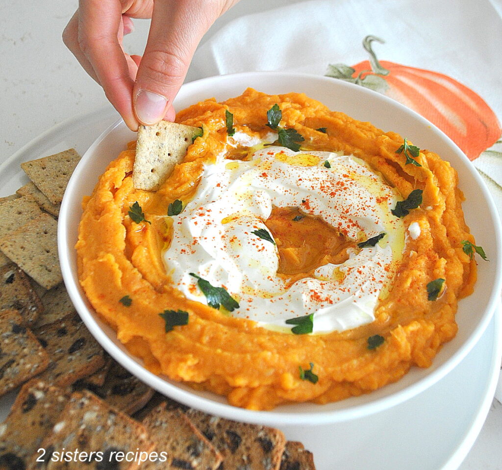 A white bowl filled with a sweet potato dip and someone is dipping with a small cracker.