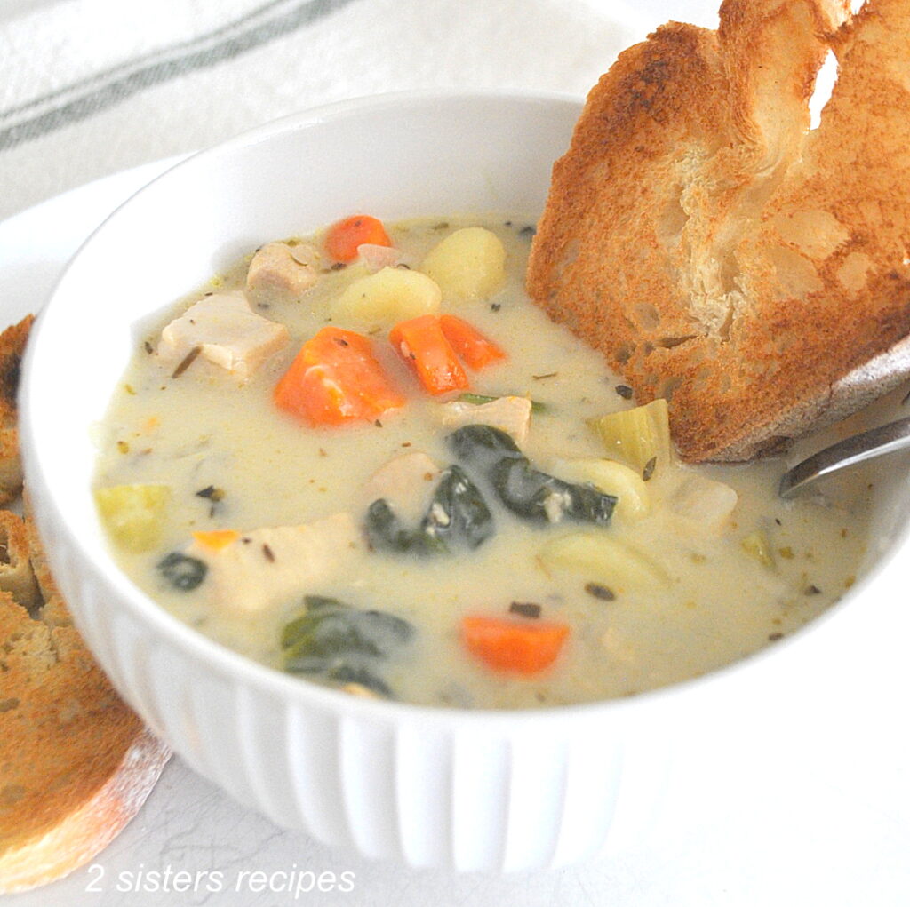 A white bowl filled with chicken gnocchi soup with veggies and a slice of toast dunk into the souo.