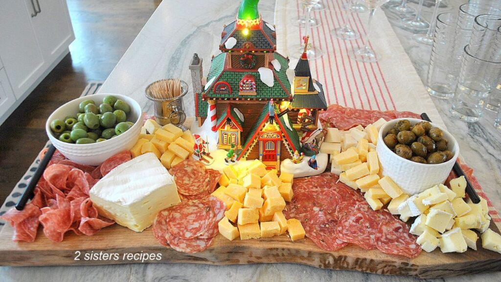 A large charcuterie board filled with a variety of cheeses, deli meats, and a Christmas house in the center.