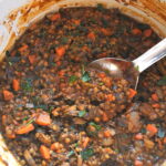 A Dutch oven pot filled with mushrooms, lentils and carrots with a large spoon inside.