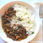A white bowl filled with a mushroom and lentil stew with a side of white rice and some grated parmesan cheese on top.