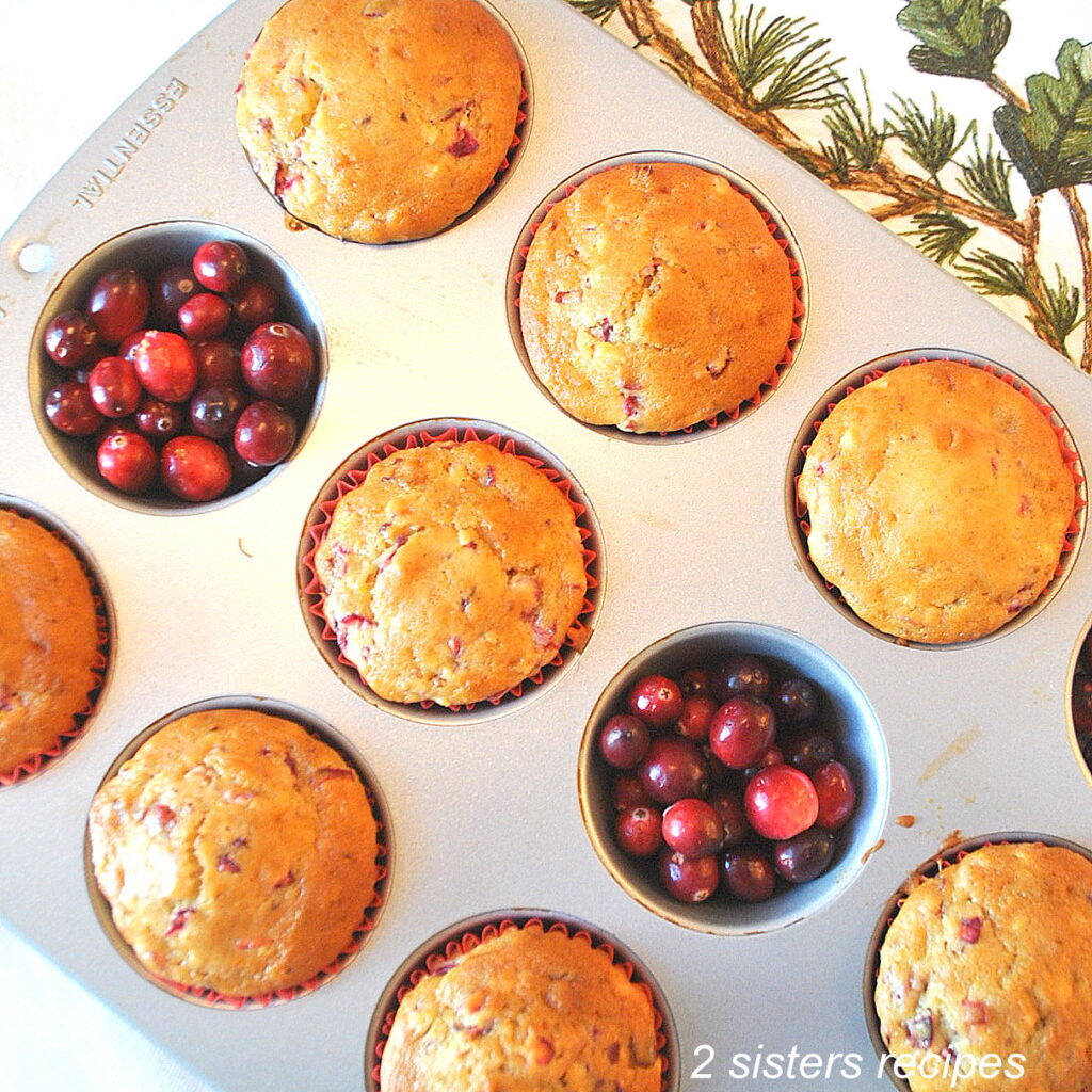 A muffin pan filled with baked muffins and fresh cranberries filled inside two muffin cups.