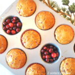A muffin pan filled with baked muffins and fresh cranberries filled inside two muffin cups.