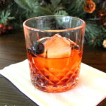 A crystal glass filled with an old fashioned cocktail, one large ice cube and a dark cherry as garnish.