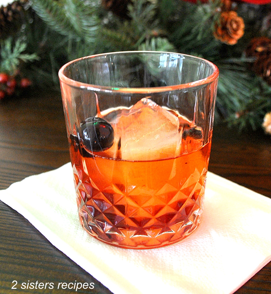 Holiday Old Fashioned Cocktail A crystal glass filled with an old fashioned cocktail, one large ice cube and a dark cherry as garnish.