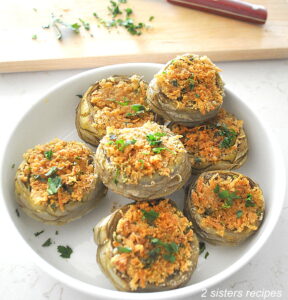 A white serving bowl filled with artichoke bottoms stuffed with bread crumb mixture and baked.