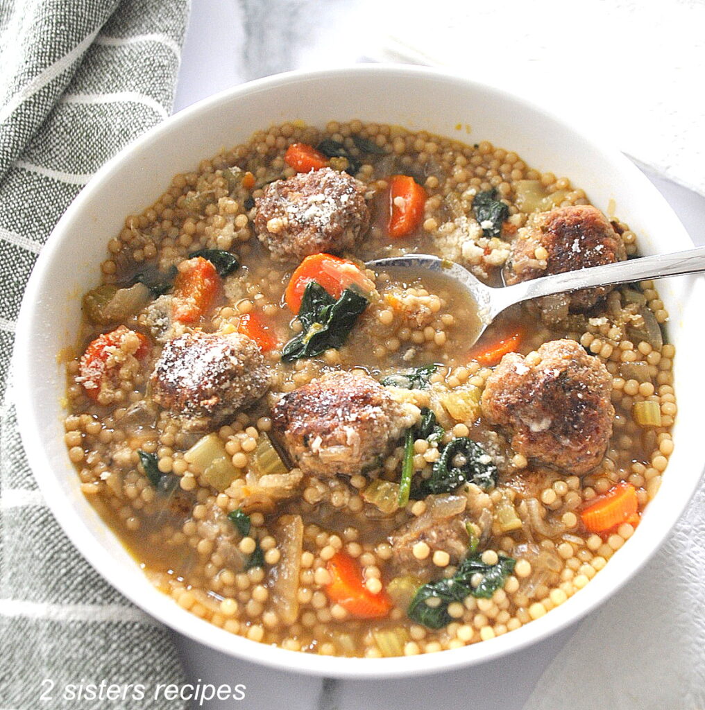 A white bowl of soup loaded with vegetables, meatballs and little round pasta. with a spoon in the bowl.