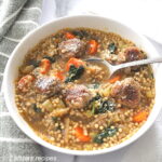A white bowl of soup loaded with vegetables, meatballs and little round pasta. with a spoon in the bowl.