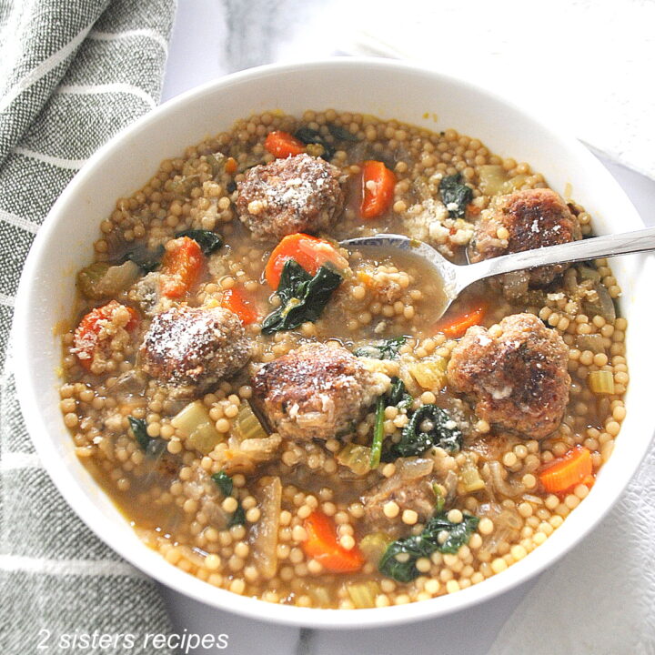 A white bowl of soup loaded with vegetables, meatballs and little round pasta. with a spoon in the bowl.
