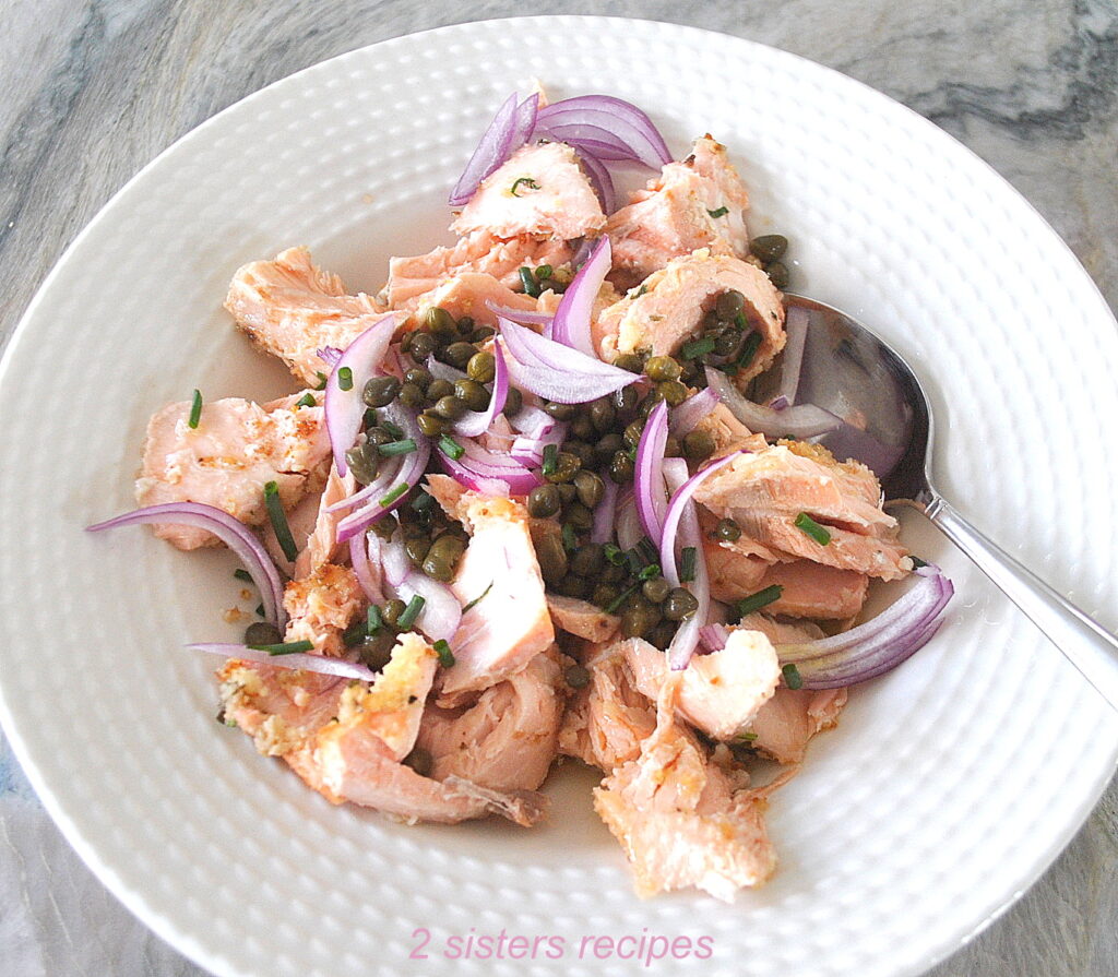 A white shallow bowl filled with chopped salmon, sliced red onion, capers and chives.