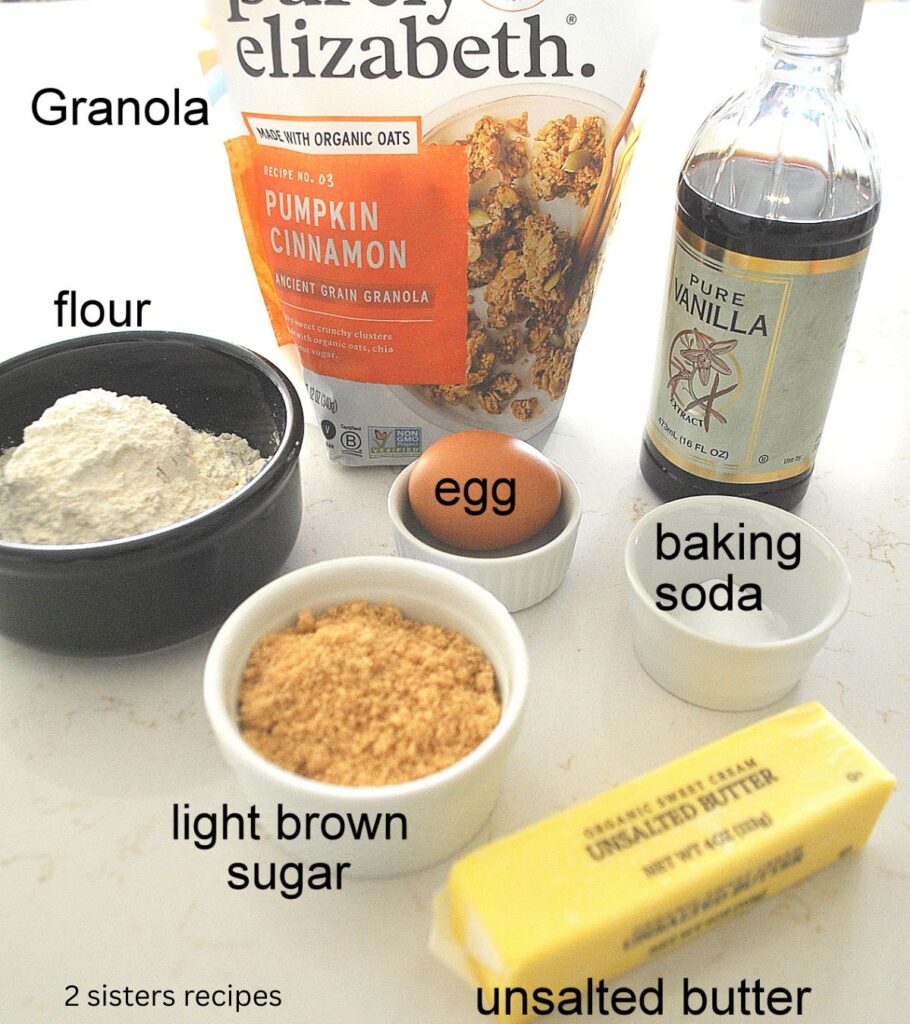 A package of granola, a stick of butter, bottle of vanilla, and bowl with flour, one egg on the counter.