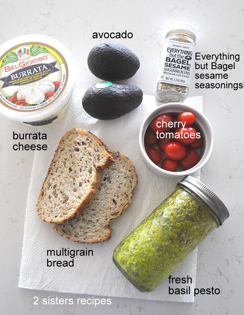 Two slices of bread, a jar of pesto sauce, and bowl of cherry tomatoes, and container of burrata cheese and 2 avocados plus a jar of seasonings on the counter.