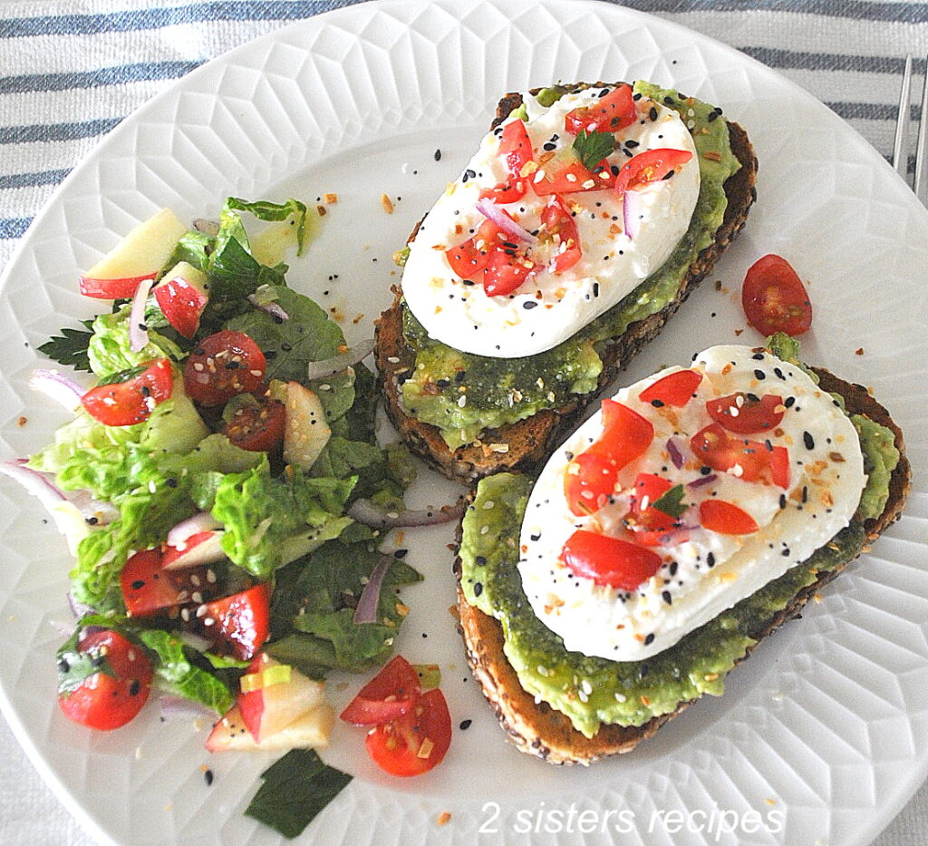 Two avocado and burrata cheese toasts on a white plate with a side salad.