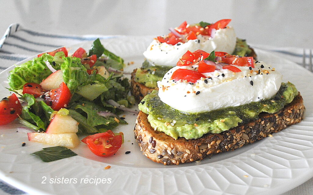 Two burrata cheese and avocado toasts served on a white dinner plate with a green salad on the side.